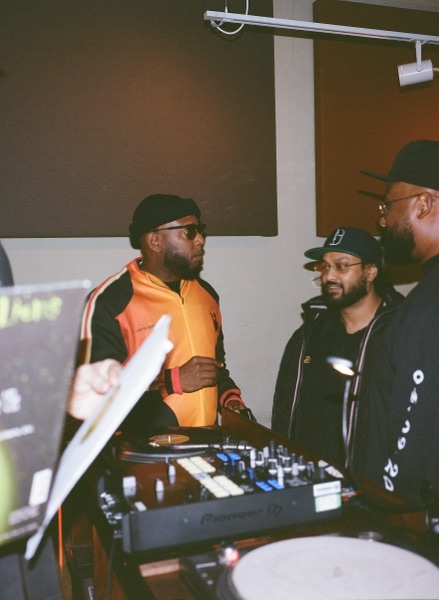 A man in an orange and black track jacket, flat cap and dark glasses stands behind a mixer holding a record sleeve. Two men in caps watch from the right of frame.