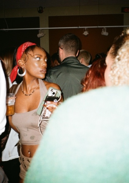 A woman in a red bandana and large hoop earrings holds a phone at chest height, looking off to one side in a busy room. A pale green blurred shoulder crosses the foreground.