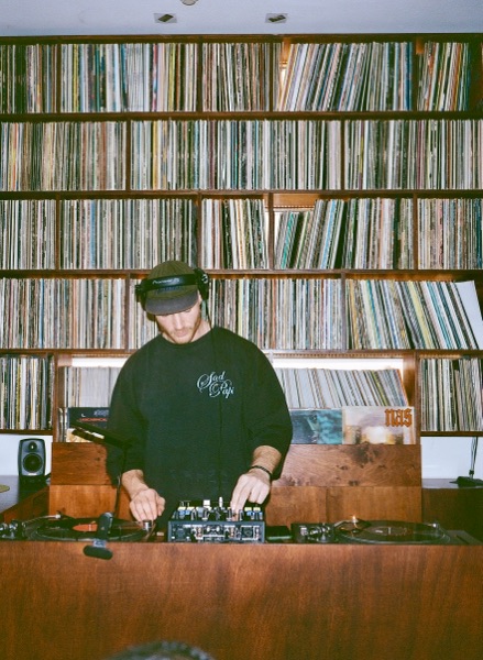 A DJ in a black sweatshirt and headphones stands at the decks, head down, in front of a floor-to-ceiling wooden shelving wall packed with vinyl.