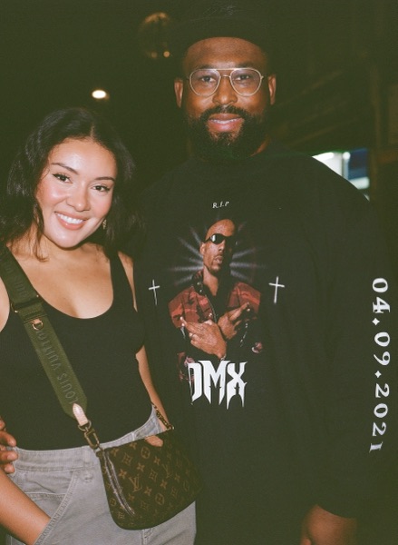 A woman in a black vest top with a monogrammed bag strap stands smiling beside a bearded man in clear-framed glasses wearing a black memorial T-shirt printed with a portrait. The film border runs down the right edge carrying a printed date stamp.