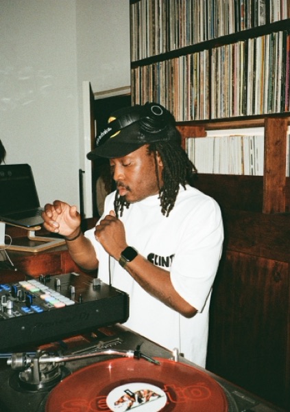 A DJ in a cap and headphones, hair in locs, works a mixer in a white T-shirt. Record shelves fill the wall behind and a red record turns on the platter.