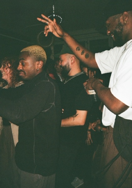 A man with bleached hair dances with his eyes closed while another reaches an arm overhead, fingers spread. A tightly packed crowd fills the rest of the frame.