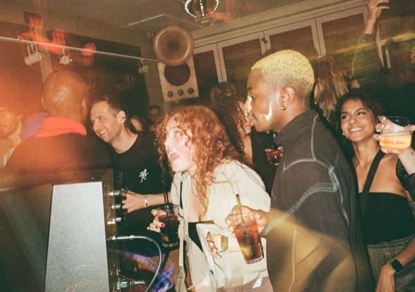 People stand at the front of a DJ booth holding drinks. A woman with curly red hair laughs mid-sentence beside a man with bleached hair. Red light streaks across the image.