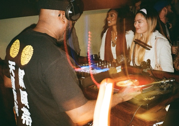A DJ photographed from behind in a black T-shirt printed with gold and white lettering, hands on the mixer. Two people laugh across the booth. A warm streak of lens flare crosses the frame.