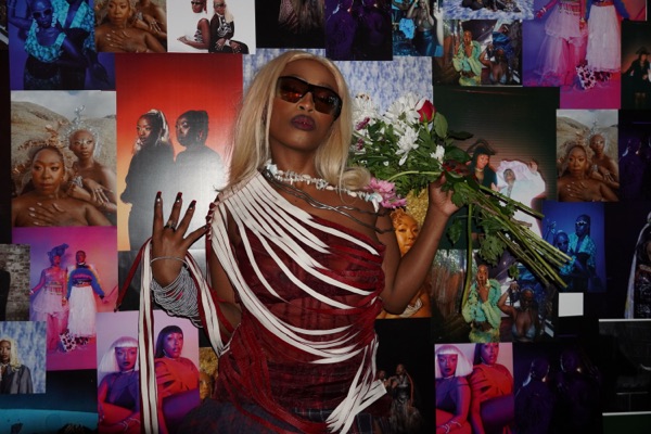 A performer in dark sunglasses and a red and white fringed dress holds a bouquet of flowers, standing in front of a wall completely covered in overlapping photographic prints.