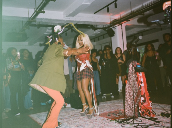 Two performers dance together on a patterned rug in the middle of a room, one in a green jacket and orange trousers, the other in a red and white fringed outfit, watched by a ring of people.