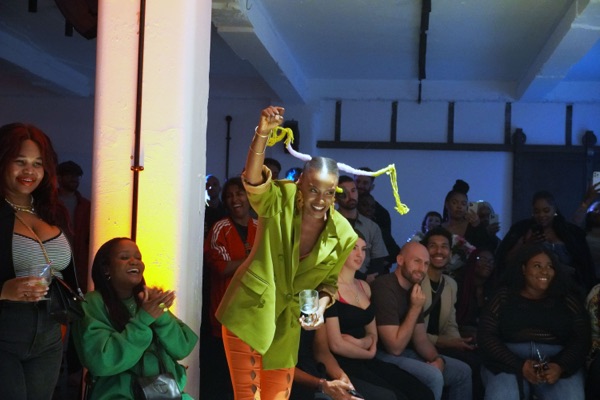 A person in a lime green suit jacket and orange trousers grins and holds something above their head, surrounded by a seated and standing crowd in a white-walled room.