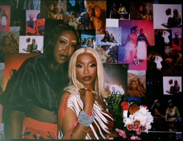 Two people pose cheek to cheek in front of a wall of overlapping photographic prints, one holding a bunch of flowers wrapped in white paper.