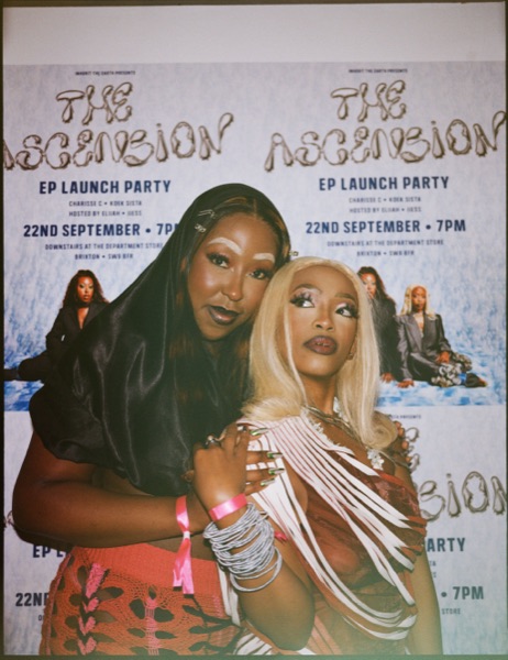 Two people pose close together in front of a wall papered with repeated printed posters. The posters carry large display lettering and several lines of smaller text.