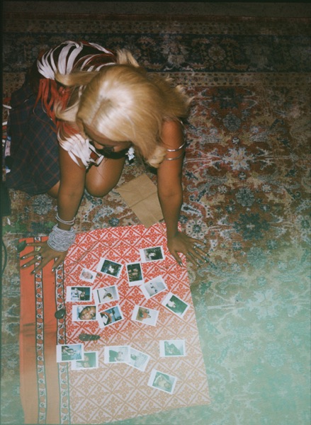 Overhead view of a person with pale hair kneeling on a patterned rug, laying out small instant photographs in rows on a red and white printed cloth.
