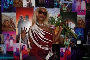 A performer in dark sunglasses and a red and white fringed dress holds a bouquet of flowers, standing in front of a wall completely covered in overlapping photographic prints.