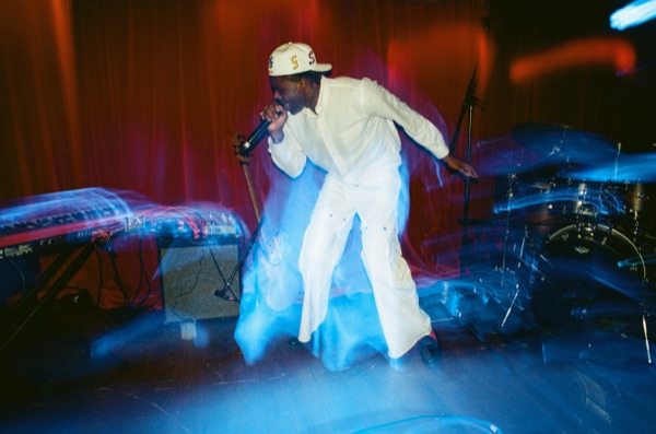 A performer in a white suit and cap leans forward singing into a microphone on a stand, caught mid-movement under blue and red stage light, drum kit to the right.