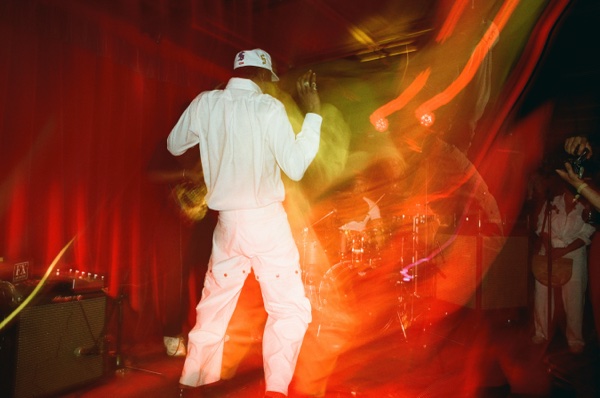 A performer in a white shirt, white trousers and a white cap stands with their back to the camera on a red-lit stage, one arm raised. Orange and yellow light trails smear across the right of the frame.