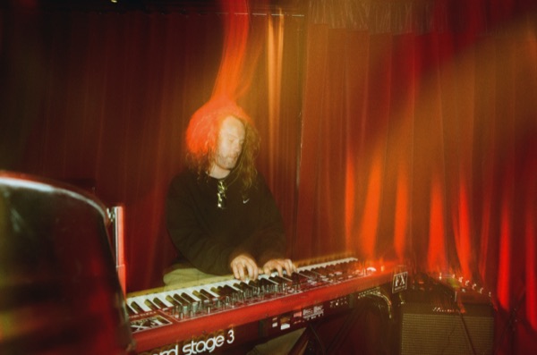 A keyboard player with long hair leans over a red stage keyboard, lit by warm light against a red curtain, motion blurring the frame.