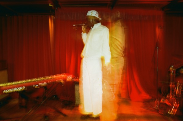 A performer in a white outfit and cap sings into a microphone in front of a red curtain, the long exposure doubling their figure into a ghost beside them.