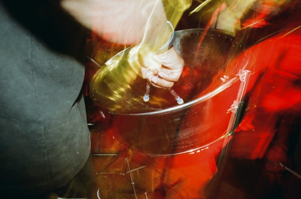 A close view of a hand gripping the rim of a large steel pan drum, the sticks blurred in motion, with red light streaking behind.