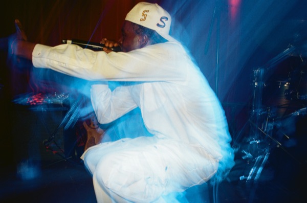 A performer in a white outfit and cap crouches low with a microphone, heavily blurred by movement, under blue stage light with a drum kit behind.