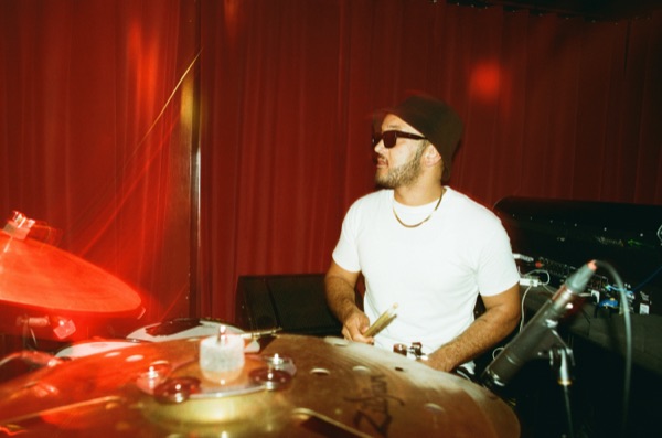 A drummer in a white T-shirt, dark cap and sunglasses sits behind a cymbal, sticks in hand, in front of a heavy red curtain.