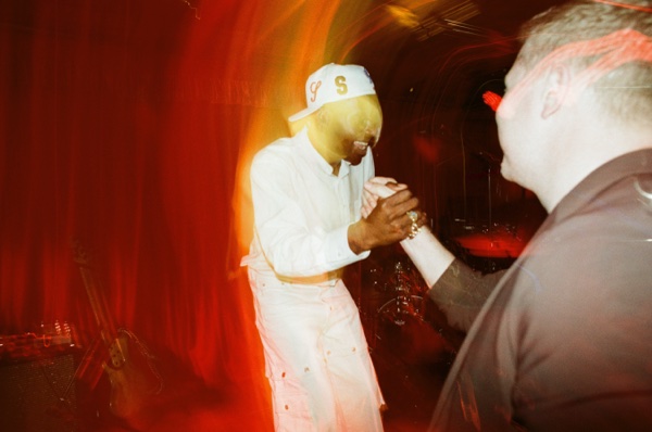 A performer dressed head to toe in white, in an embroidered cap, leans forward to clasp hands with someone at the front of the crowd. A long orange light trail curves through the frame.