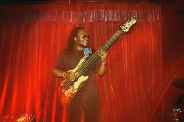 A bass player in a dark red T-shirt and sunglasses, locs falling past the shoulder, smiles while playing a five-string bass in front of a red stage curtain.