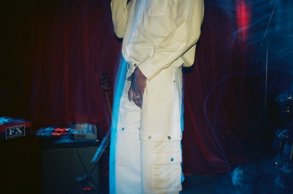A cropped view of someone standing in a cream denim jacket and matching wide trousers against a dark red curtain, a pale blue light streak running down beside them. An amplifier and a pedalboard sit at the left.