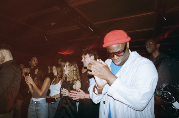 A man in a red beanie, sunglasses and white jacket claps his hands in a dark room, a crowd behind him holding drinks.