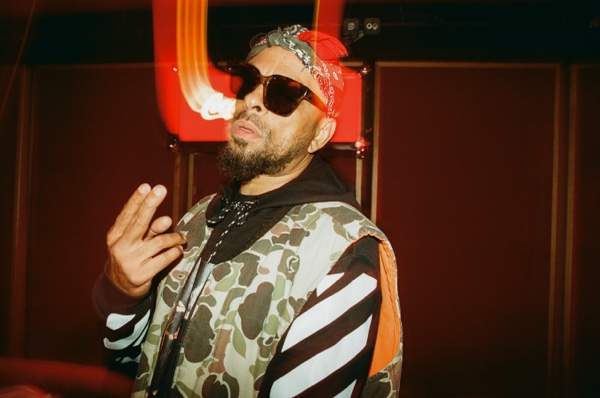 A man in a red patterned bandana and dark sunglasses, in a camouflage and diagonal-striped jacket, holds up a two-finger sign. A red neon tube glows on the wall behind him.