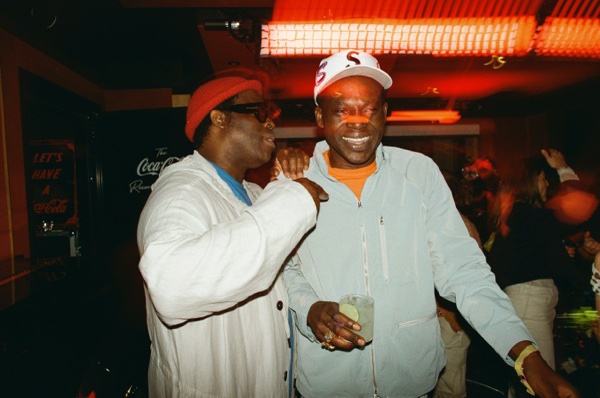 Two men stand close together in a dark room under a red strip light, one in a red beanie and a loose white jacket with an arm across the other's shoulder, the other in a white cap and pale blue zip-up jacket holding a drink.