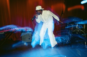 A performer in a white suit and cap leans forward singing into a microphone on a stand, caught mid-movement under blue and red stage light, drum kit to the right.