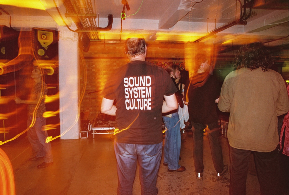 A man photographed from behind wearing a black T-shirt printed with three lines of white text, standing in a brick-walled basement room among a small crowd. Streaks of orange light trail across the frame.